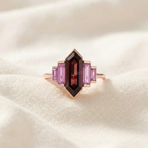 May include: A rose gold ring featuring a central elongated hexagonal dark red garnet gemstone flanked by two smaller baguette-cut light purple sapphires on each side, set in a symmetrical arrangement on a thin polished rose gold metal band.