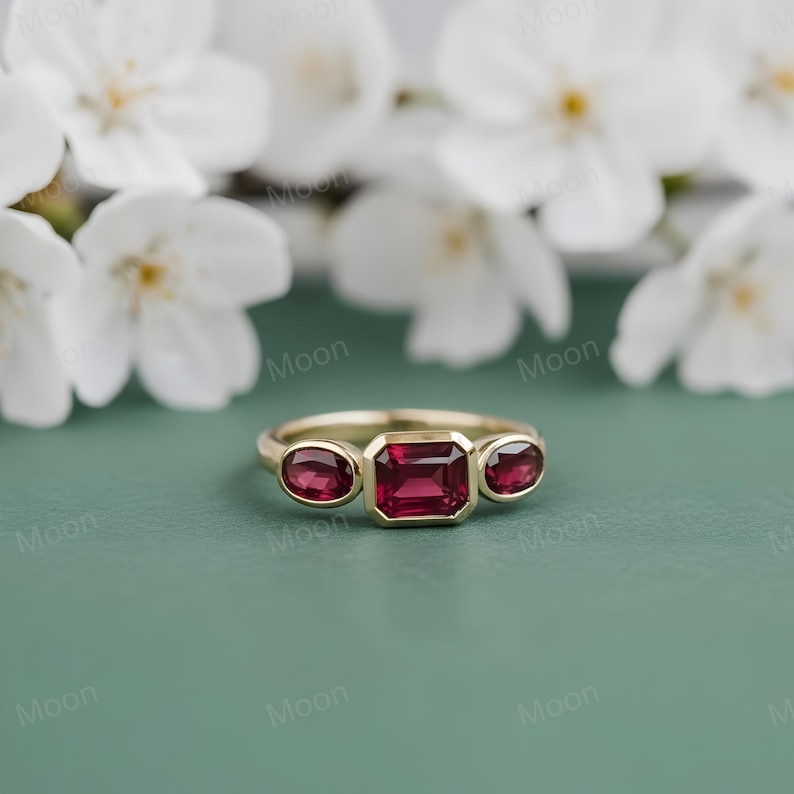 May include: A gold ring with three gemstones. The center stone is a rectangular, deep red gemstone, flanked by two oval-shaped, smaller red gemstones. The ring is set against a soft green background with white flowers.