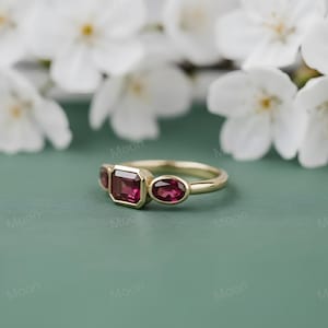 May include: A gold ring with three gemstones. The center stone is a rectangular cut, flanked by two oval-cut stones. All stones are a deep red color. The ring is set against a soft green background with white flowers.