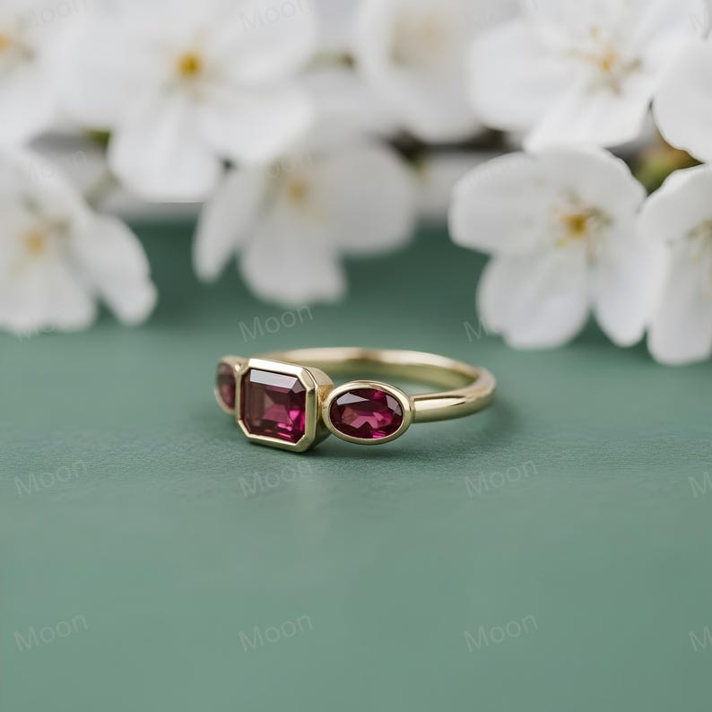 May include: A gold ring with three gemstones. The center stone is a rectangular cut, flanked by two oval-cut stones. All stones are a deep red color. The ring is set against a soft green background with white flowers.