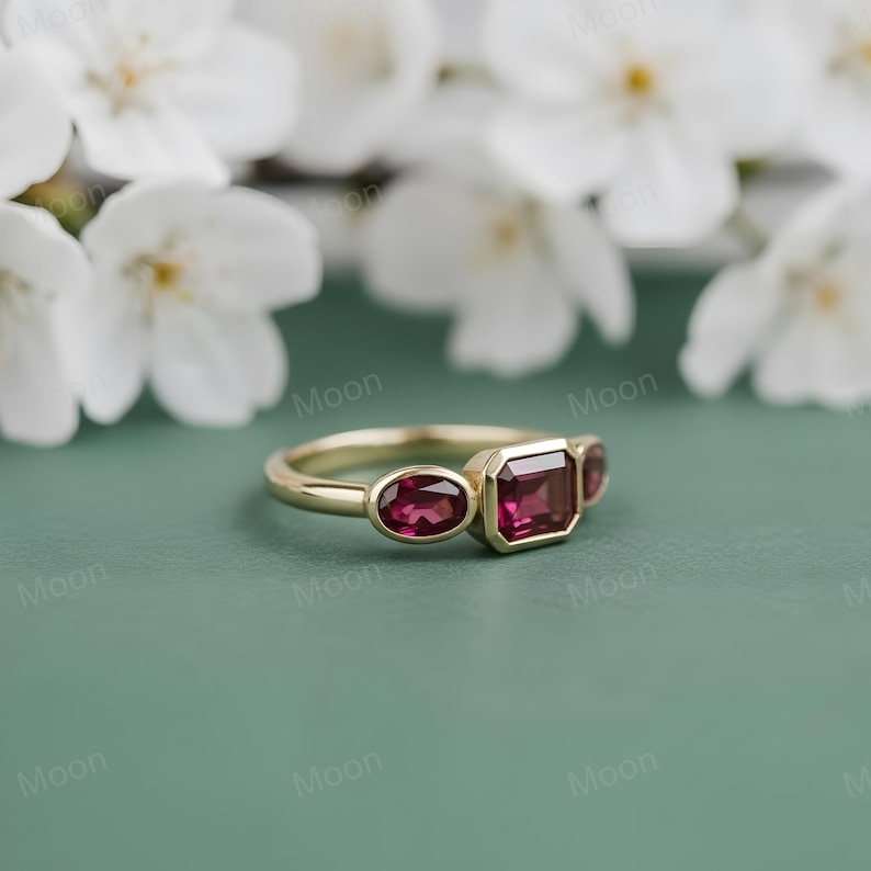 May include: A gold ring with three red gemstones. The center stone is an emerald cut, flanked by two oval-shaped stones. The ring is set against a green background with white flowers.