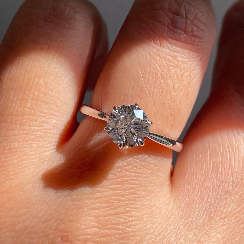 May include: A close-up of a silver ring with a large, round, clear gemstone. The ring is set with six prongs, and the band is thin and polished. The gemstone sparkles in the light.