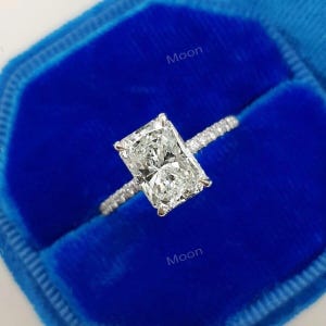 May include: A diamond ring with a rectangular cut stone, presented in a blue velvet box. The band is embellished with smaller diamonds. The ring is set against a deep blue backdrop, creating a striking contrast.