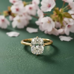 May include: A gold ring with a large, rectangular, clear gemstone. The ring is set against a green background with blurred white flowers in the background. The ring is a classic design.