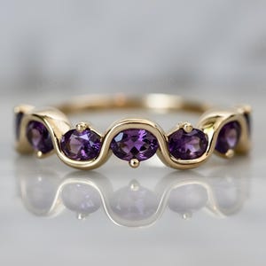 Pode incluir: Um anel de ouro com uma série de pedras preciosas roxas ovais. As pedras são colocadas num padrão ondulado. A banda do anel é de ouro polido e as pedras são de um roxo profundo. Joia.