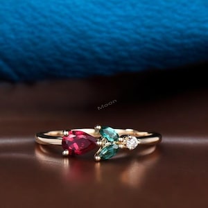 以下が含まれることがあります： ゴールドのリング。洋梨型の深い赤色の宝石、2つの緑色の葉の形をした石、小さな丸い透明な石が特徴です。 リングはぼやけた背景にあります。