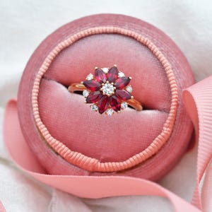 May include: A gold ring with a red ruby flower design. The ring is set in a pink velvet ring box.