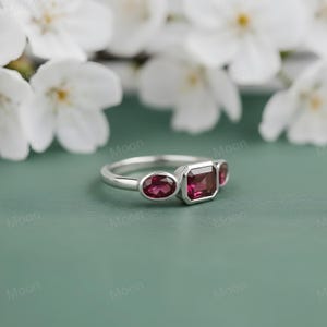 May include: A silver ring with three red gemstones. The ring features an oval-shaped stone on each side of a rectangular center stone. The ring is set against a green background with white flowers.