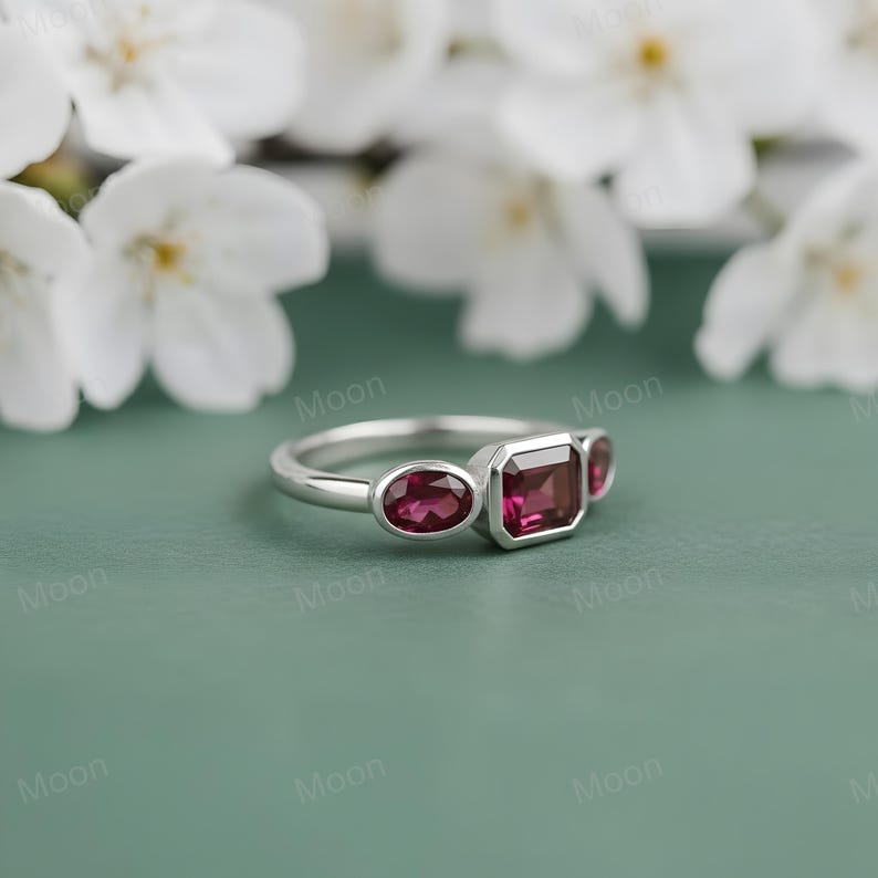 May include: A silver ring with three red gemstones. The ring features an oval-shaped stone on each side of a rectangular center stone. The ring is set against a green background with white flowers.