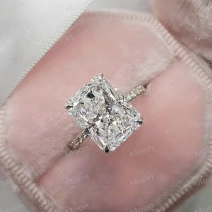 Op de afbeelding: Een diamanten ring met een rechthoekige slijpvorm, gepresenteerd in een roze fluwelen doosje. De ring heeft een grote, heldere diamant en een band versierd met kleinere diamanten. De zetting is zachtroze, wat de schittering van de ring versterkt.