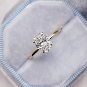 Puede incluir: Un anillo de compromiso de oro con un diamante de talla ovalada. El diamante está engastado con cuatro garras y se asienta sobre una fina banda de oro. El anillo se muestra en una caja de terciopelo azul claro.