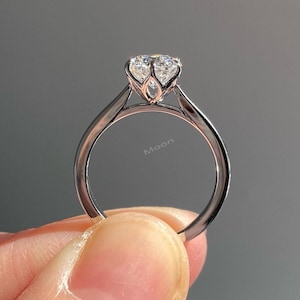 May include: A silver engagement ring with a large, clear gemstone. The ring has a delicate, openwork setting and a slender band. The word "Moon" is visible on the band. The ring is held by a person.