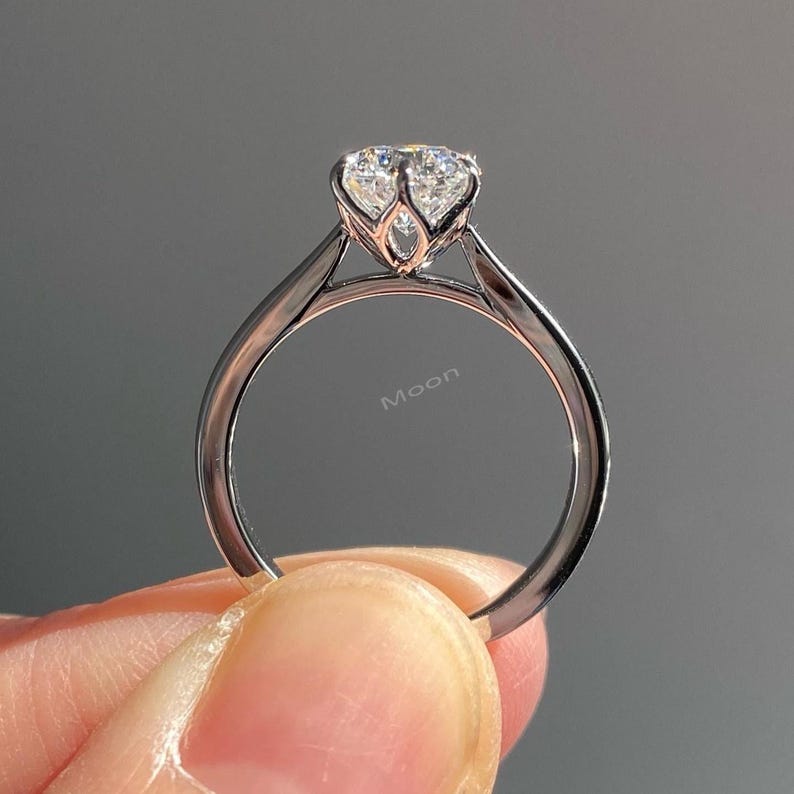 May include: A silver engagement ring with a large, clear gemstone. The ring has a delicate, openwork setting and a slender band. The word "Moon" is visible on the band. The ring is held by a person.