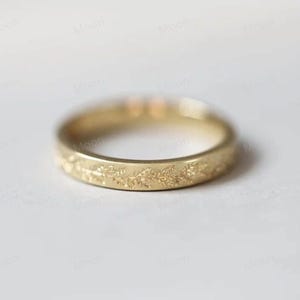 Könnte beinhalten: Ein goldfarbener Ring mit einem floralen Muster, das um das Band geätzt ist. Der Ring hat eine glatte, polierte Oberfläche und ein zartes Design. Der Ring ist wahrscheinlich aus Gold oder einem goldfarbenen Metall gefertigt. Der Ring ist ein Schmuckstück.