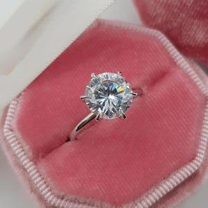 Puede incluir: Un anillo de compromiso solitario con un gran diamante de talla redonda engastado en una banda plateada. El anillo se muestra en un joyero de terciopelo rosa con una cinta blanca.