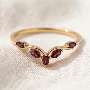 May include: A gold V-shaped ring featuring five marquise-cut deep red garnet gemstones arranged in a chevron pattern along the band. The polished gold band curves to follow the shape of the stones, creating a delicate and symmetrical jewellery piece.