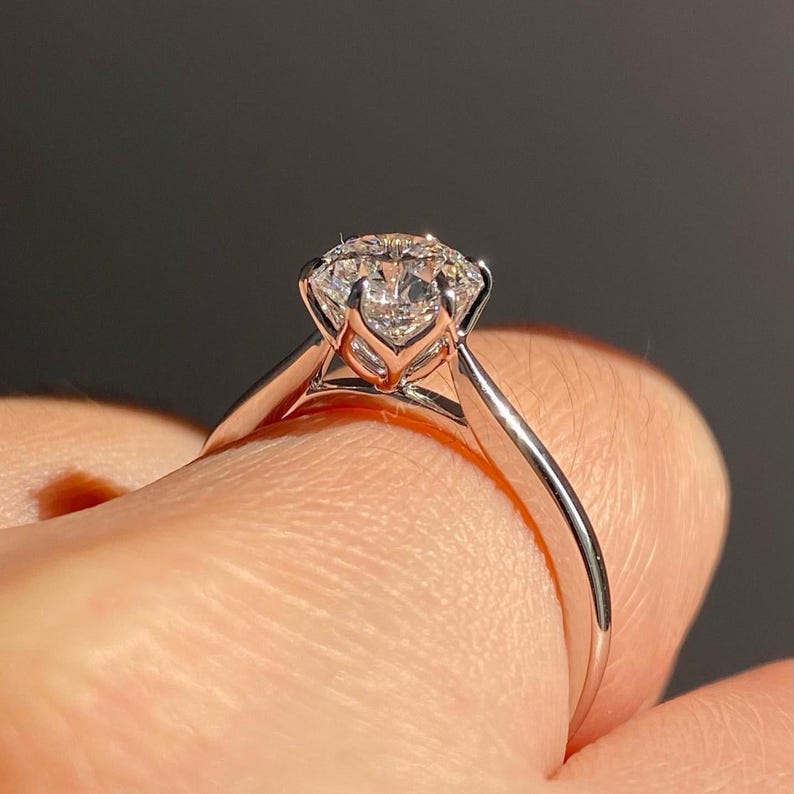 May include: A close-up of a solitaire engagement ring. The ring features a large, round-cut diamond set in a classic, four-prong setting. The band is a polished, silver-colored metal, likely platinum or white gold. The ring is held up against a dark background.