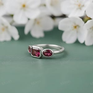 May include: A silver ring featuring three gemstones. The center stone is a rectangular cut, flanked by two oval-cut stones. All three stones are a deep red color. The ring is set against a soft green background with white flowers.