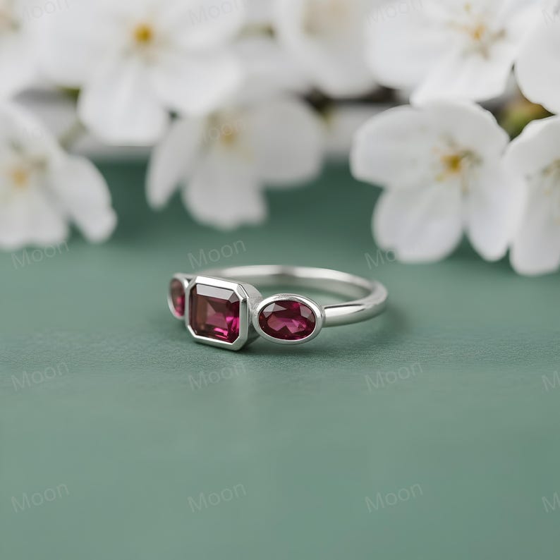 May include: A silver ring featuring three gemstones. The center stone is a rectangular cut, flanked by two oval-cut stones. All three stones are a deep red color. The ring is set against a soft green background with white flowers.