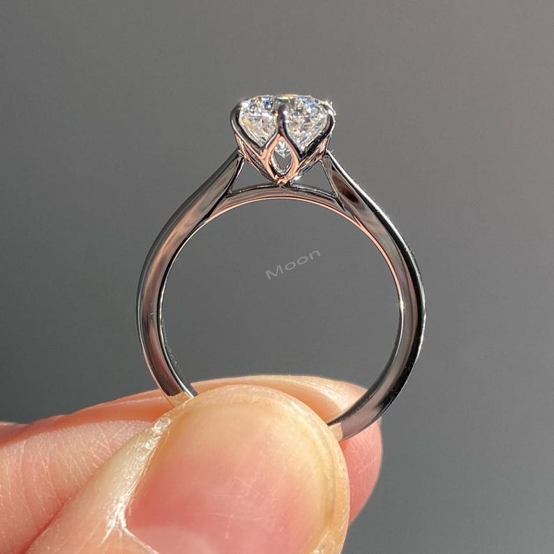 May include: A close-up of a silver ring with a large, clear gemstone. The ring has a delicate, minimalist design with a slender band. The gemstone is set in a prong setting, and the word "Moon" is engraved on the band.