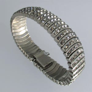Può includere: Bracciale color argento con un design flessibile e a coste. Il bracciale è adornato con più file di pietre preziose chiare e scintillanti. La chiusura è un design semplice e sicuro. Il gioiello è esposto su uno sfondo chiaro.