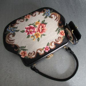 May include: A black handbag and a matching small coin purse featuring needlepoint embroidery with a central floral bouquet of pink, red, and yellow flowers surrounded by a cream-colored background and a black border with decorative scrollwork.