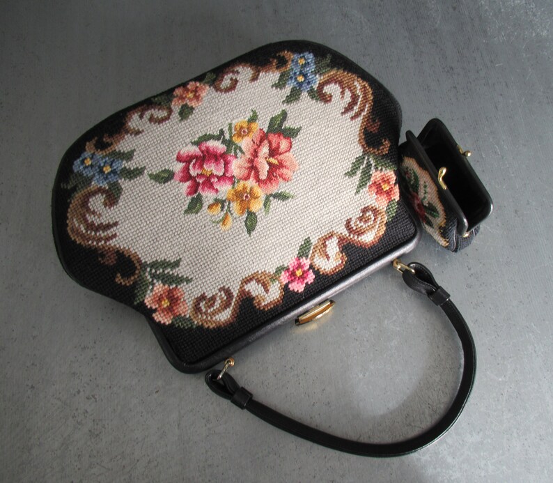 May include: A black handbag and a matching small coin purse featuring needlepoint embroidery with a central floral bouquet of pink, red, and yellow flowers surrounded by a cream-colored background and a black border with decorative scrollwork.