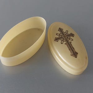 May include: An open, oval-shaped, off-white trinket box with a matching lid. The lid features a brown cross design. The box is likely made of plastic or a similar material. The background is a solid, light gray color.
