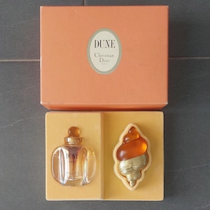 Może przedstawiać: Zestaw perfum Dune od Christian Dior. Zestaw zawiera mały, przezroczysty flakon perfum ze złotą nakrętką i dekoracyjny element w kształcie muszli. Prezentowany w brzoskwiniowym pudełku z logo Dune.
