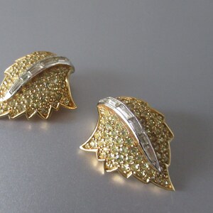 May include: A pair of gold tone leaf-shaped earrings with clear and yellow rhinestones. The earrings have a silver tone accent in the center.