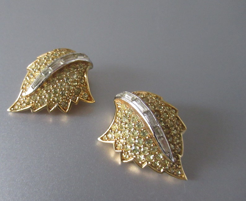 May include: A pair of gold tone leaf-shaped earrings with clear and yellow rhinestones. The earrings have a silver tone accent in the center.