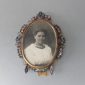 Può includere: Un ritratto ovale antico in una cornice decorativa. La cornice è dorata con abbellimenti ornati di accenti bianchi, blu e rosa. Il ritratto è una fotografia in bianco e nero di una donna con un vestito bianco e una collana di perle.