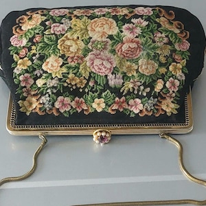 Peut inclure: Une pochette noire ornée d'un motif floral en point de tapisserie avec des fleurs roses, jaunes et vertes. Le sac possède un cadre en métal doré, une fine bandoulière en chaîne dorée et un fermoir décoratif agrémenté de perles et d'une pierre.