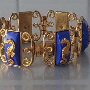 Op de afbeelding: Goudkleurige armband met rechthoekige blauwe emaille panelen met gouden zeepaardontwerpen. De armband heeft een sierlijk, opengewerkt ontwerp met verbindingsschakels. Een sieraad in vintage stijl.