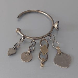 Op de afbeelding: Een zilverkleurige open armband met een donkere ronde steenzetting en vier bungelende bedels. De bedels bestaan uit een getextureerde bladvorm, een effen cirkel, een ruitvorm en een grotere platte vierkante hanger, allemaal verbonden met schakels.