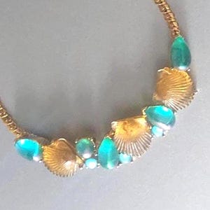 Könnte beinhalten: Eine goldfarbene Kettenhalskette mit einer geschwungenen Anordnung aus metallisch goldenen Jakobsmuscheln, durchsetzt mit glatten, tropfenförmigen blaugrünen und türkisfarbenen Perlen, die einen halbkreisförmigen Blickfang bilden.