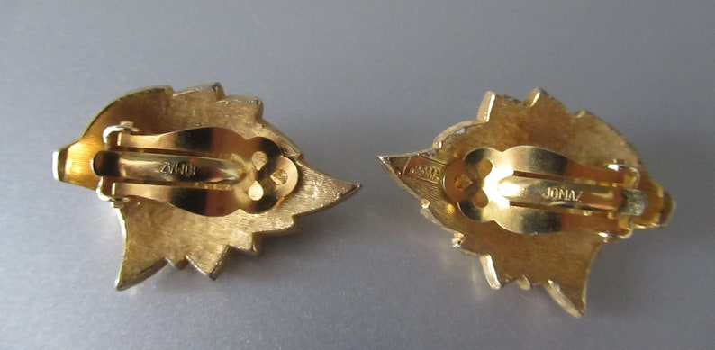 May include: A pair of gold-tone clip-on earrings with a leaf design. The back of the earrings are marked "JONAV".