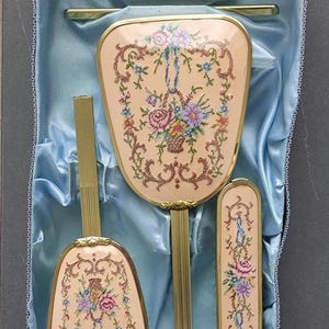 Op de afbeelding: Een vintage toilettafelset in een blauw satijn gevoerde doos. De set bevat een handspiegel, een borstel en een kam, allemaal met een crèmekleurige achtergrond en bloemenborduurwerk. De handvatten zijn goudkleurig.
