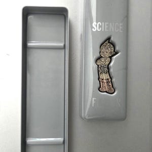 May include: A rectangular gray metal pencil case with a removable lid. The lid features the word SCIENCE printed in white, the word FICTION partially visible below, and a central embossed graphic of a cartoon character with spiky hair.