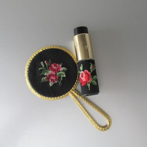 May include: A gold-coloured hand mirror with a black fabric backing featuring an embroidered floral design of red roses. A small, black and gold perfume bottle with a floral design is also pictured.