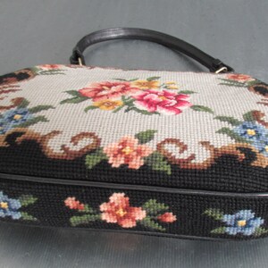 May include: An oval-shaped handbag featuring a needlepoint floral design. The top panel displays a light gray background with pink, yellow, and blue flowers, while the sides are black with floral accents. It includes a single black leather handle.