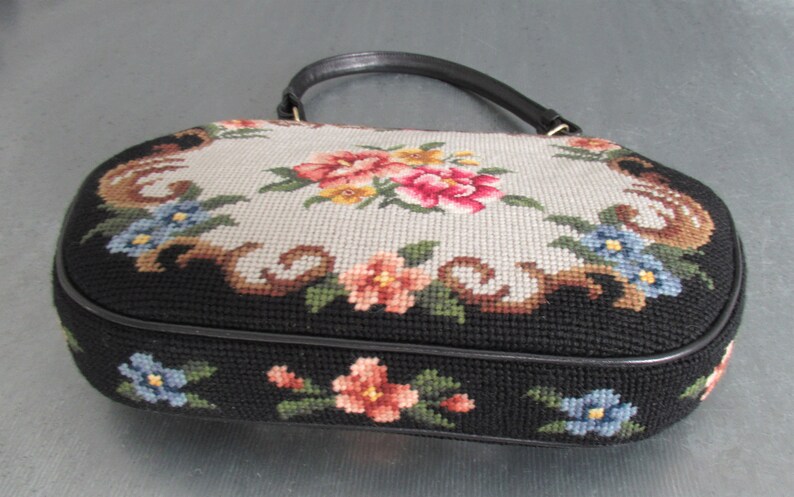 May include: An oval-shaped handbag featuring a needlepoint floral design. The top panel displays a light gray background with pink, yellow, and blue flowers, while the sides are black with floral accents. It includes a single black leather handle.