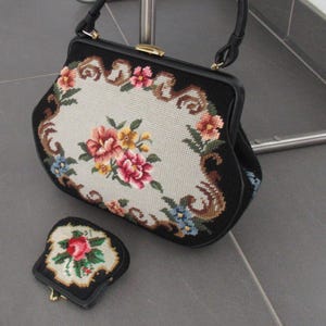 May include: A black handbag and a matching coin purse featuring needlepoint floral patterns on a cream background. The handbag has a black frame, a single handle, and a gold-tone metal clasp. The smaller coin purse also has a gold-tone metal clasp.