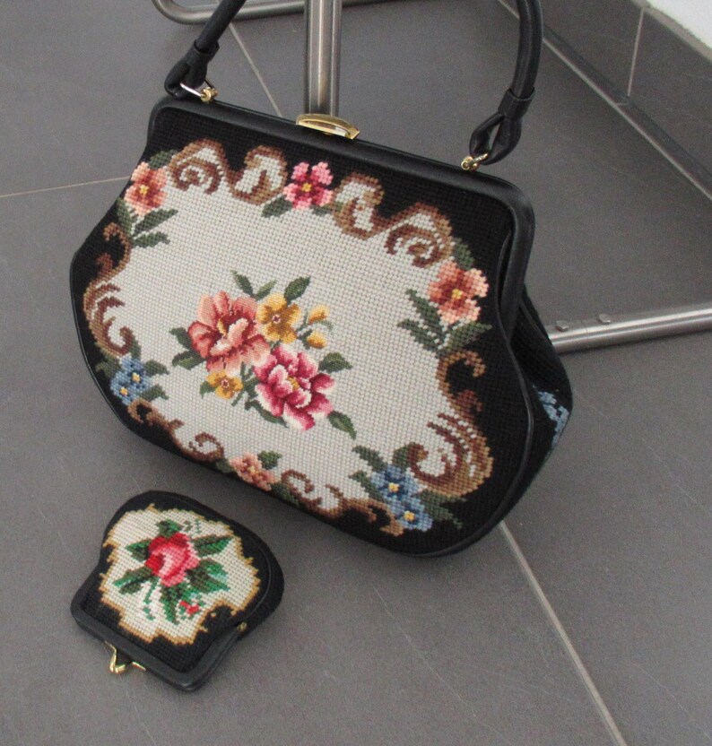 May include: A black handbag and a matching coin purse featuring needlepoint floral patterns on a cream background. The handbag has a black frame, a single handle, and a gold-tone metal clasp. The smaller coin purse also has a gold-tone metal clasp.
