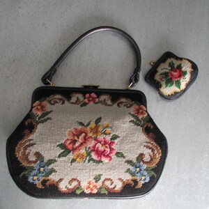 May include: A black handbag with a single handle and a matching small coin purse. Both items feature a needlepoint floral design with pink, yellow, and blue flowers on a light gray background, framed by a black border and gold-tone hardware.