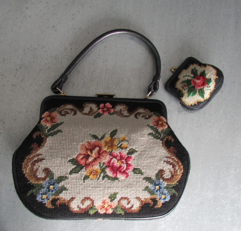 May include: A black handbag with a single handle and a matching small coin purse. Both items feature a needlepoint floral design with pink, yellow, and blue flowers on a light gray background, framed by a black border and gold-tone hardware.