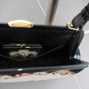 May include: A black handbag with a needlepoint floral design on the exterior, featuring a gold-tone metal clasp and a black strap. The interior is lined in black fabric and includes a small matching needlepoint coin purse with a gold-tone frame.