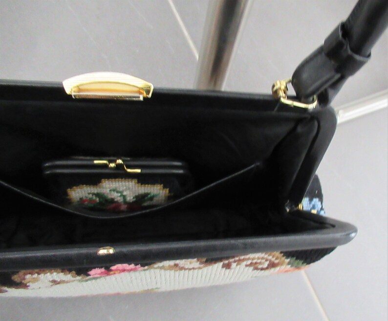 May include: A black handbag with a needlepoint floral design on the exterior, featuring a gold-tone metal clasp and a black strap. The interior is lined in black fabric and includes a small matching needlepoint coin purse with a gold-tone frame.