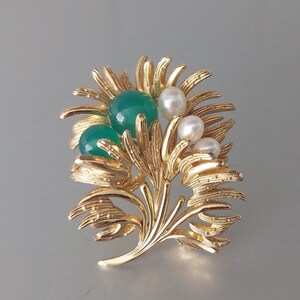 Könnte beinhalten: Eine goldfarbene Brosche mit einem stilisierten floralen Design. Die Brosche hat drei grüne Edelsteine und drei weiße Perlen.