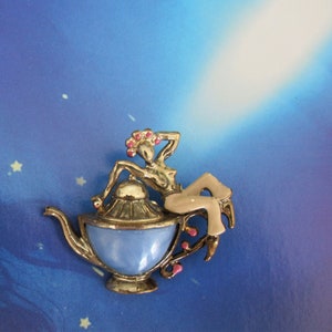 May include: A gold-toned metal brooch featuring a stylized figure reclining on a blue teapot. The figure has a tan body and pink floral accents. The teapot has a light blue body and gold trim.